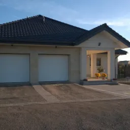 Dom jednorodzinny z elewacją wykończoną kamieniem, dwoma białymi bramami garażowymi i nowo wyłożonym podjazdem z kostki brukowej w odcieniach szarości.