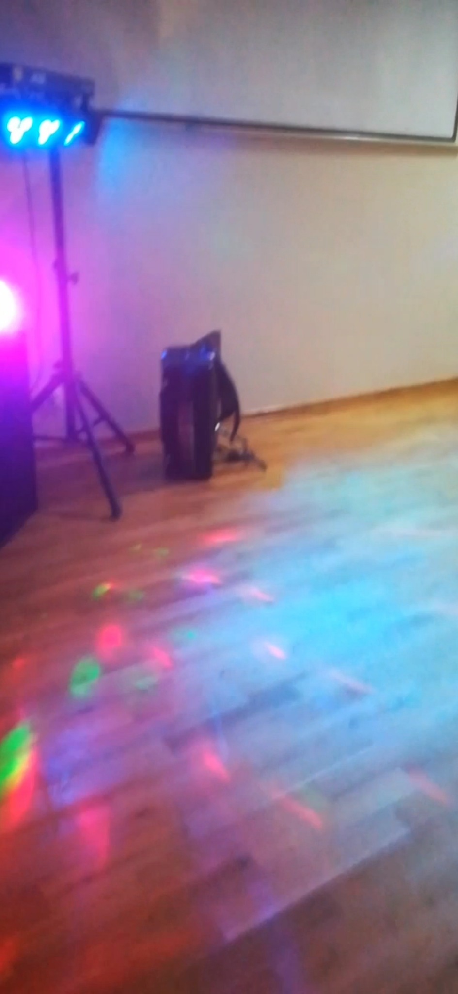 Sprzęt oświetleniowy na statywie i kolumna głośnikowa rzucające kolorowe światła na parkiet, w tle biała ściana.