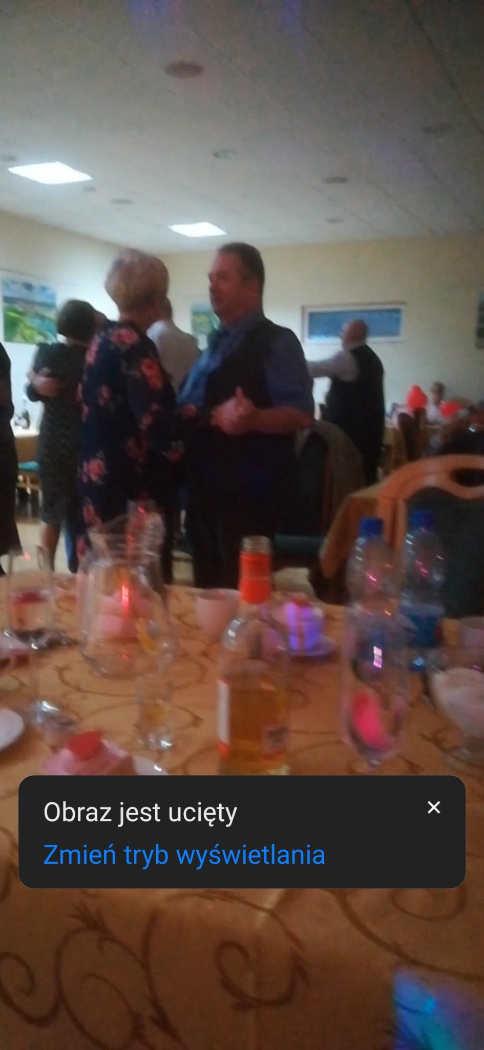 Para tańczy na sali balowej, na pierwszym planie zastawiony stół z butelkami i dzbankiem z napojami.