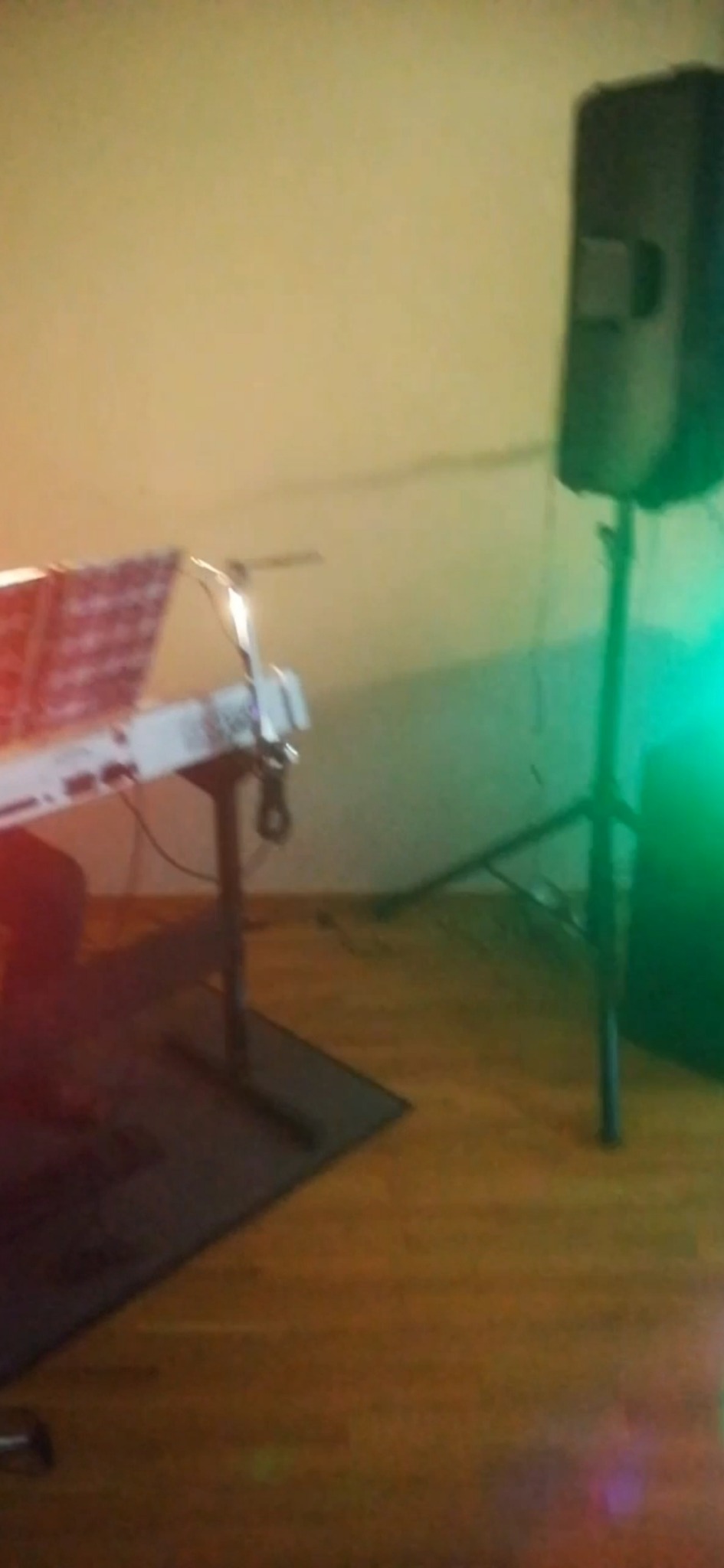 Rozstawiony keyboard z nutami i lampką, obok kolumna głośnikowa na statywie, widok z boku w słabym oświetleniu.