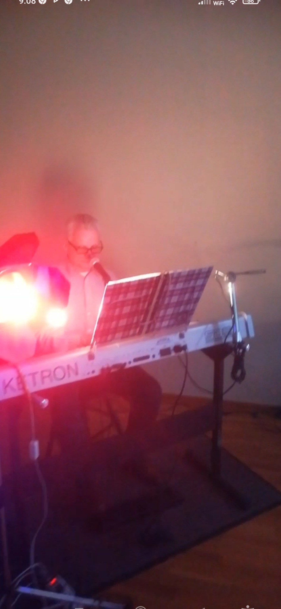 Mężczyzna w okularach gra na keyboardzie Ketron w czerwonym oświetleniu scenicznym, czytając nuty z pulpitu.