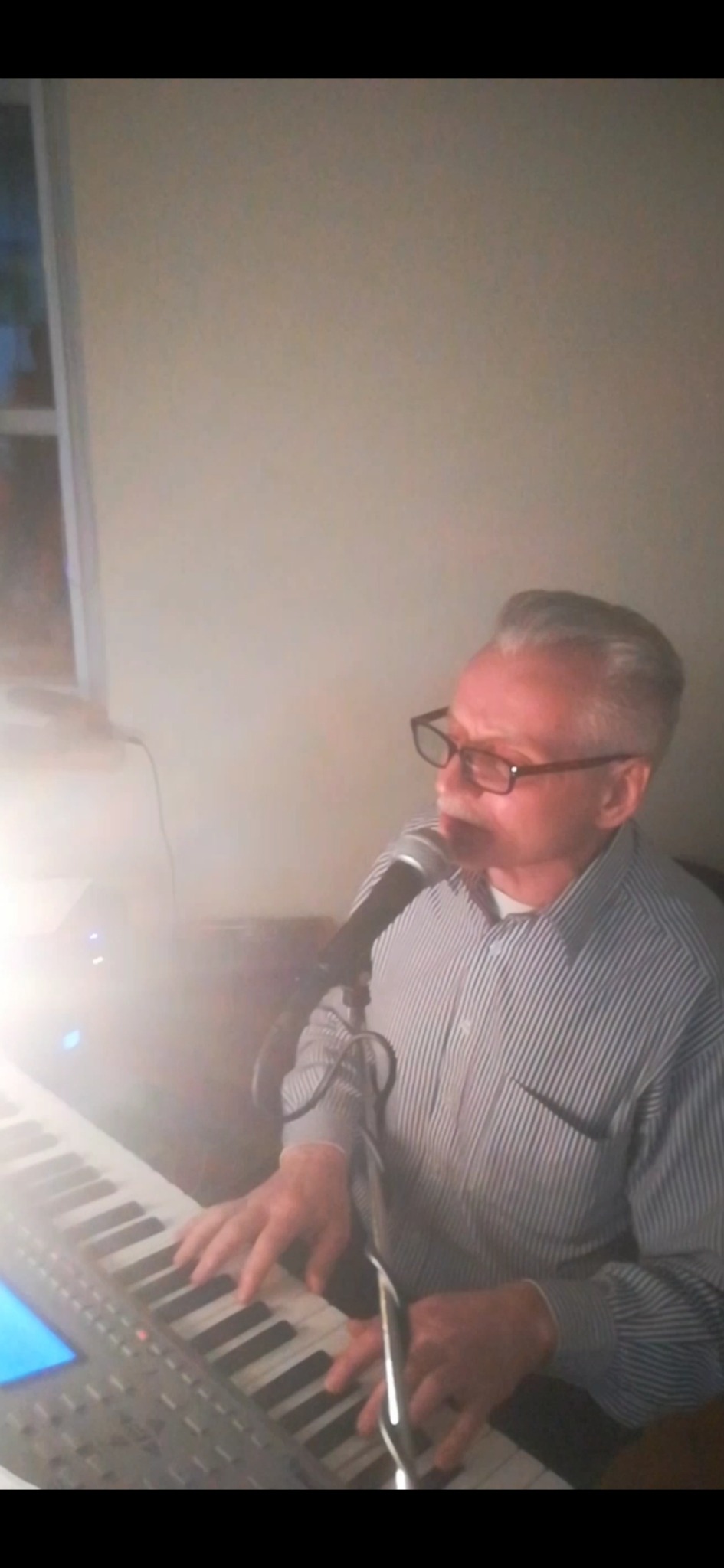 Mężczyzna w okularach i koszuli w paski śpiewa do mikrofonu i gra na keyboardzie.