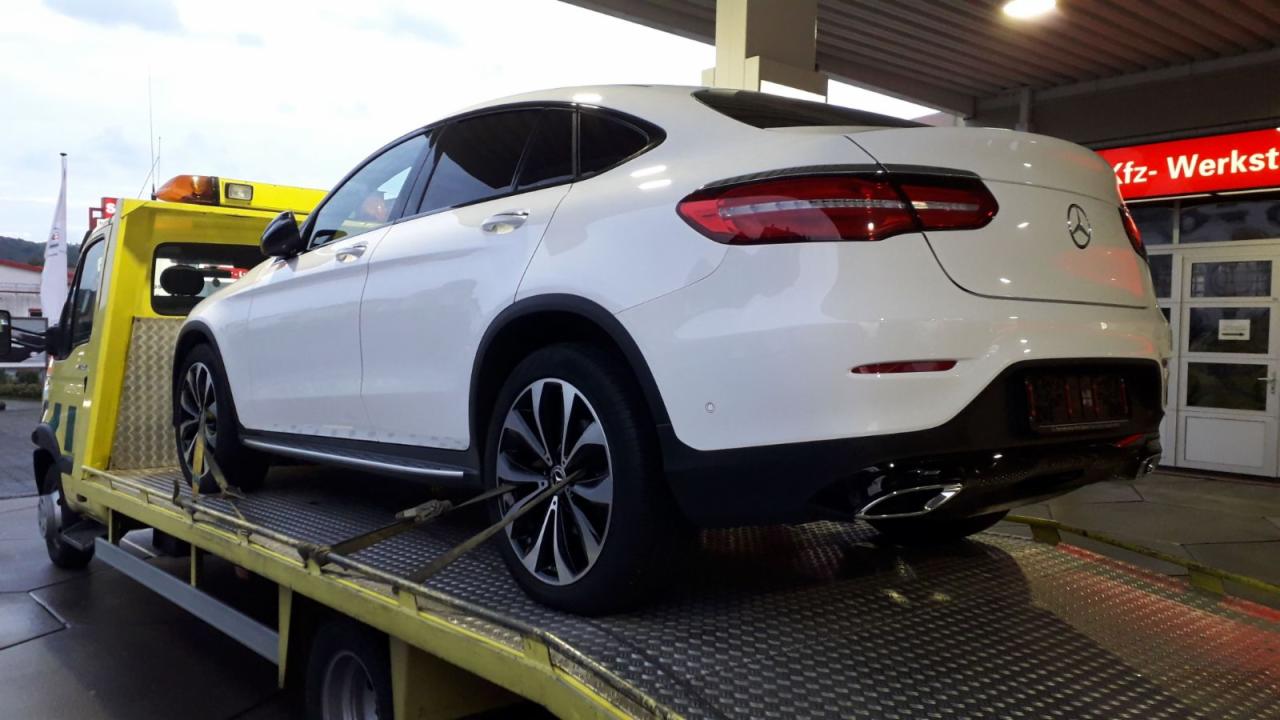 Biały Mercedes-Benz GLC Coupe zabezpieczony pasami na żółtej platformie pojazdu pomocy drogowej przed warsztatem samochodowym.