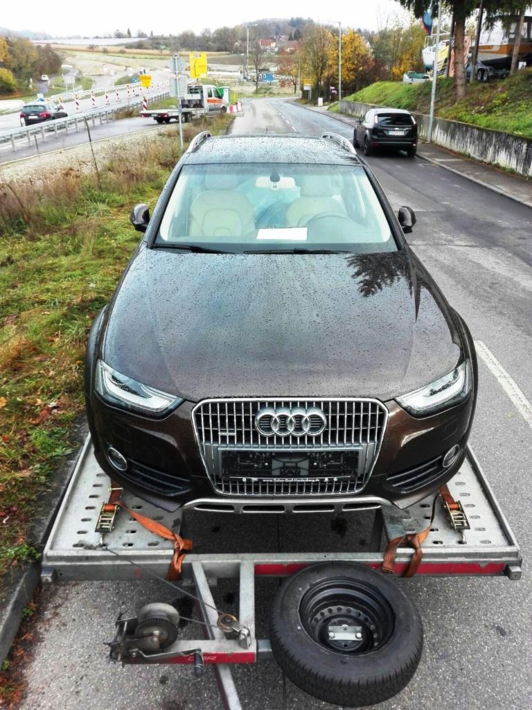 Brązowe Audi Allroad na metalowej platformie przyczepy, zabezpieczone pomarańczowymi pasami, z widocznym kołem zapasowym i wciągarką linową, na tle drogi i zieleni.