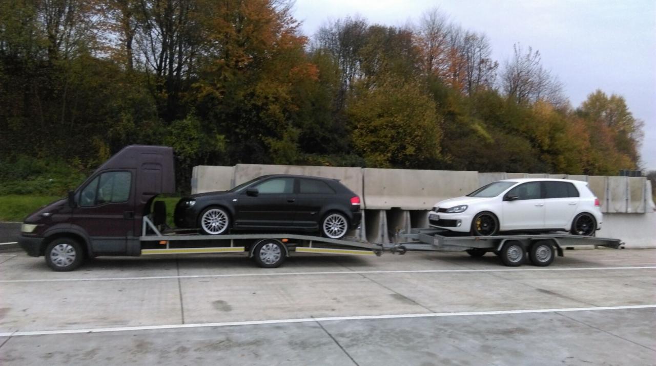 Ciemnobordowa laweta z przyczepą przewozi dwa samochody osobowe: czarny Audi A3 i biały Volkswagen Golf na tle betonowych barier i jesiennego lasu.