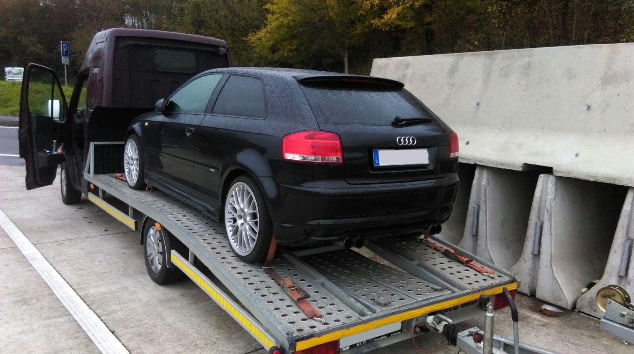 Czarny samochód Audi A3 zabezpieczony pasami na platformie lawety podczas transportu autostradą, widoczne betonowe bariery.