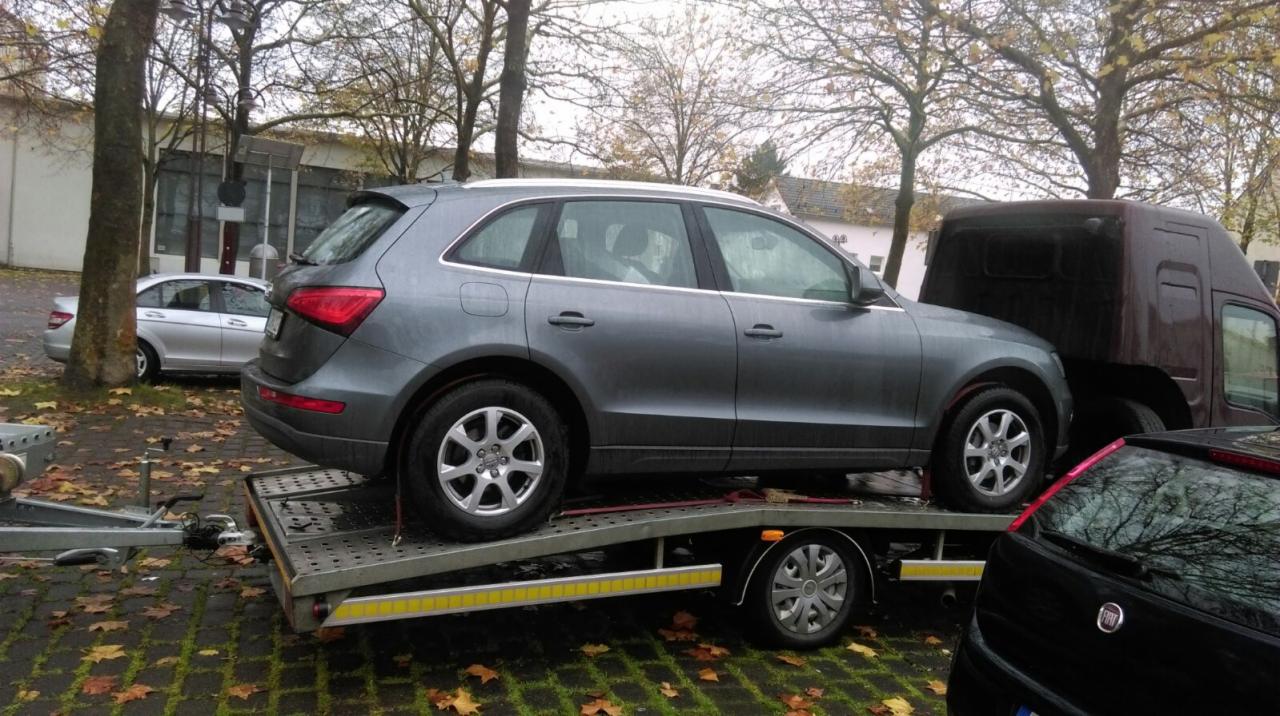 Szary samochód osobowy marki Audi Q5 zabezpieczony pasami na platformie przyczepy samochodowej, obok stoi brązowy samochód dostawczy, w tle budynek i drzewa.