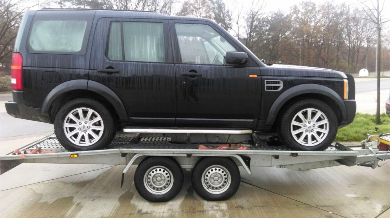 Czarny samochód terenowy marki Land Rover Discovery na metalowej platformie lawety, widok z boku, mokra nawierzchnia.