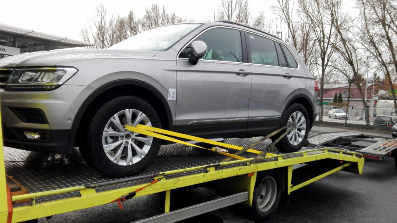 Srebrny SUV marki Volkswagen zabezpieczony żółtymi pasami transportowymi na żółtej platformie lawety w trakcie załadunku, widok z boku na tle drzew i budynków.