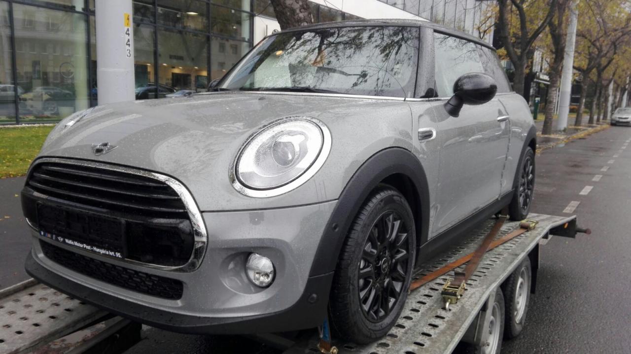 Szary Mini Cooper na metalowej platformie lawety, zabezpieczony brązowymi pasami transportowymi, widok z przodu na tle miejskiej ulicy w pochmurny dzień.
