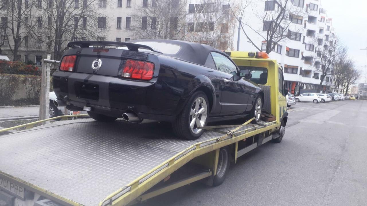 Czarny kabriolet Ford Mustang z ciemnymi pasami na żółtej lawecie na tle miejskiego krajobrazu z blokami mieszkalnymi i drzewami bez liści.