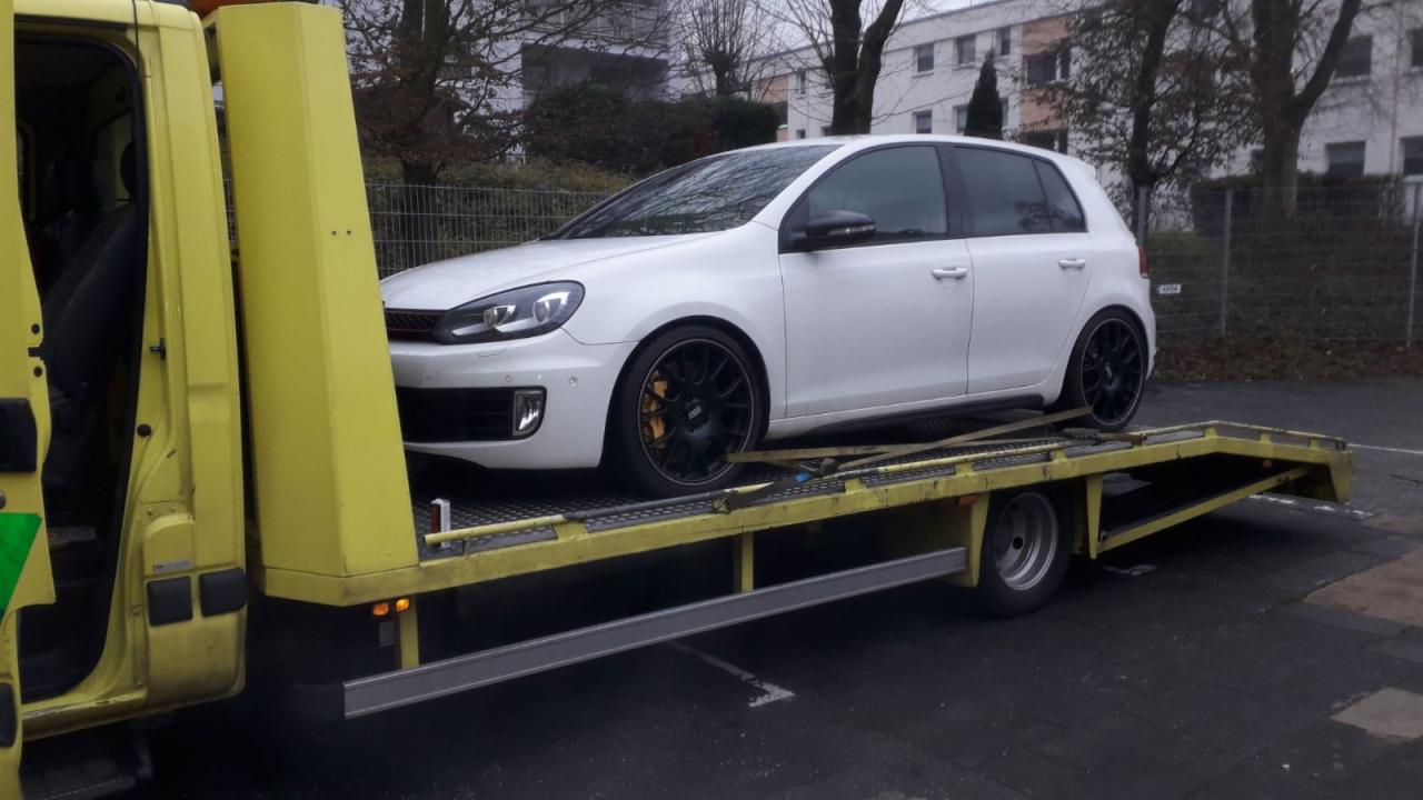 Biały Volkswagen Golf VI na żółtej lawecie, widoczne czarne felgi i żółte zaciski hamulcowe, tło stanowią drzewa i budynek mieszkalny.