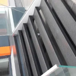 Zewnętrzne schody z ciemnego kamienia z szklaną balustradą, widok z g&oacute;ry, nowoczesna architektura budynku z pomarańczowymi elementami.