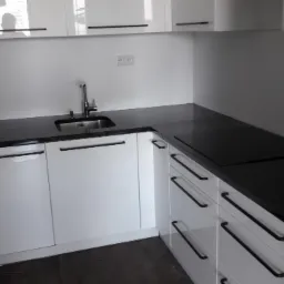 Nowoczesna kuchnia z białymi szafkami i czarnym blatem, minimalistyczne uchwyty, widoczny zlew i płyta indukcyjna, oświetlenie sufitowe odbijające się w blacie.