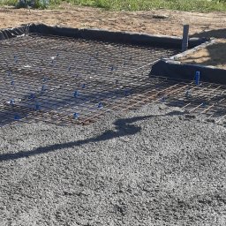 Fabryka Domu - Zalewanie betonem zbrojenia na gruncie, widoczne plastikowe podkładki dystansowe pod siatką zbrojeniową i czarna folia izolacyjna na obrzeżach wykopu.