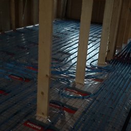 Fabryka Domu - Wodne ogrzewanie podłogowe w trakcie montażu w domu szkieletowym w Gdańsku, widoczne niebieskie rurki na folii z siatką, drewniane słupki konstrukcyjne.