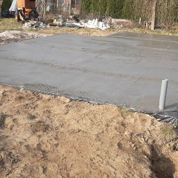 Fabryka Domu - Świeżo wylana, gładka płyta fundamentowa na czarnej folii, wystające rury kanalizacyjne. W tle narzędzia budowlane, ziemia i trawa.