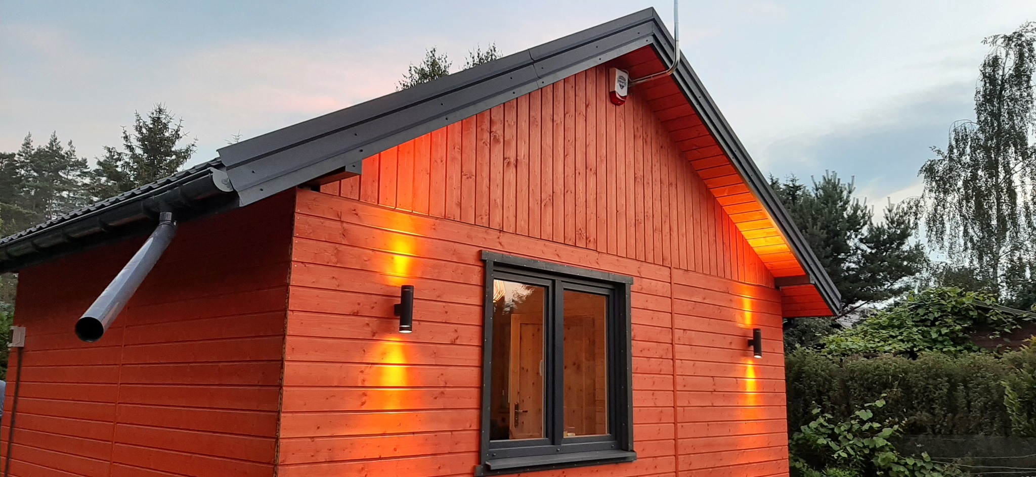 Czerwony budynek z drewnianą elewacją, oświetlony dwoma zewnętrznymi lampami ściennymi, z czarną stolarką okienną i dachem, widoczny system rynnowy i czujnik alarmowy.