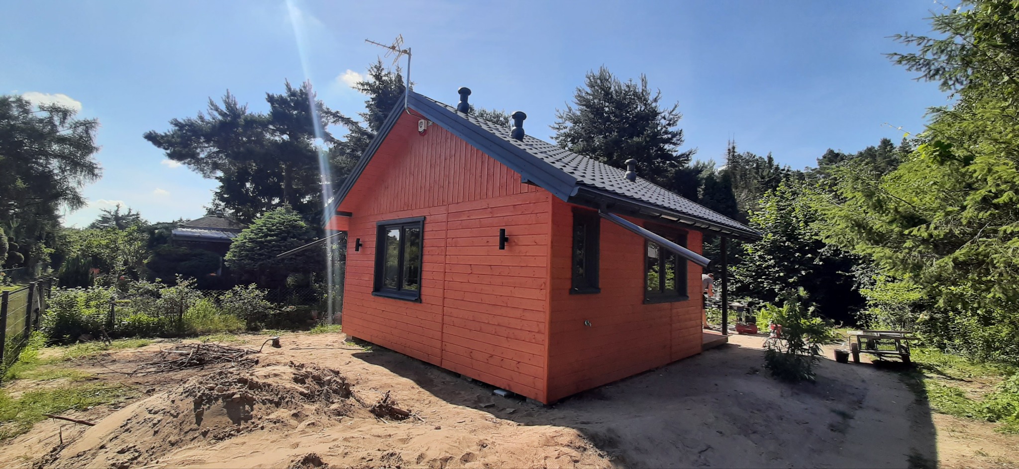 Pomarańczowy domek z drewnianych bali z ciemnoszarą dachówką, czarnymi ramami okiennymi i dwiema lampami na ścianie, otoczony zielenią i stertą piasku.