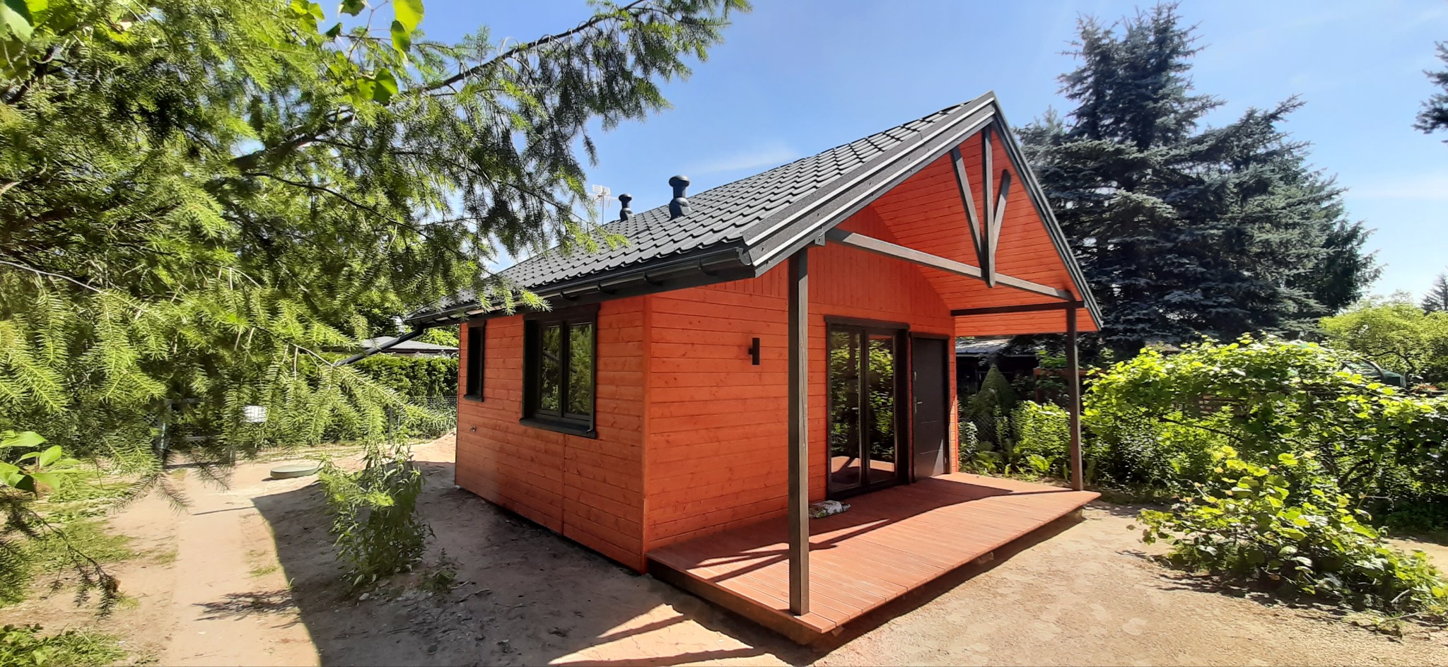Pomarańczowy domek letniskowy z ciemną stolarką okienną i zadaszonym tarasem, otoczony zielenią drzew i krzewów w słoneczny dzień.