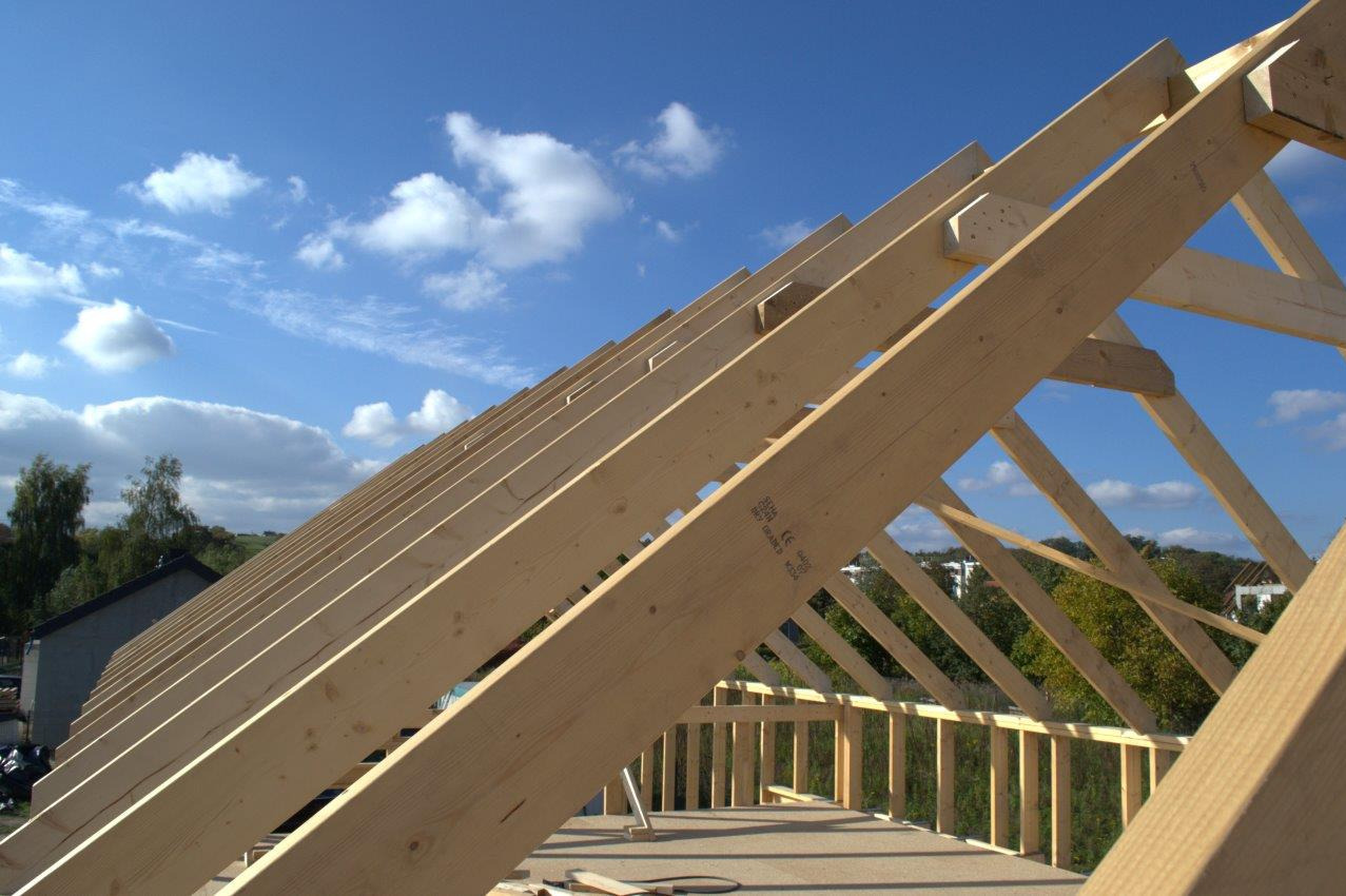 Konstrukcja drewniana dachu w trakcie budowy, widoczne elementy więźby dachowej na tle błękitnego nieba z chmurami.