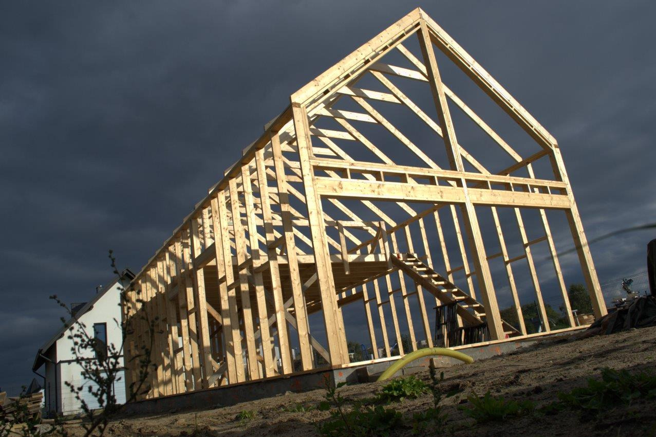 Konstrukcja domu w technologii szkieletowej w trakcie budowy, widoczne drewniane elementy nośne na tle ciemnego, burzowego nieba; w tle widoczny fragment gotowego domu.