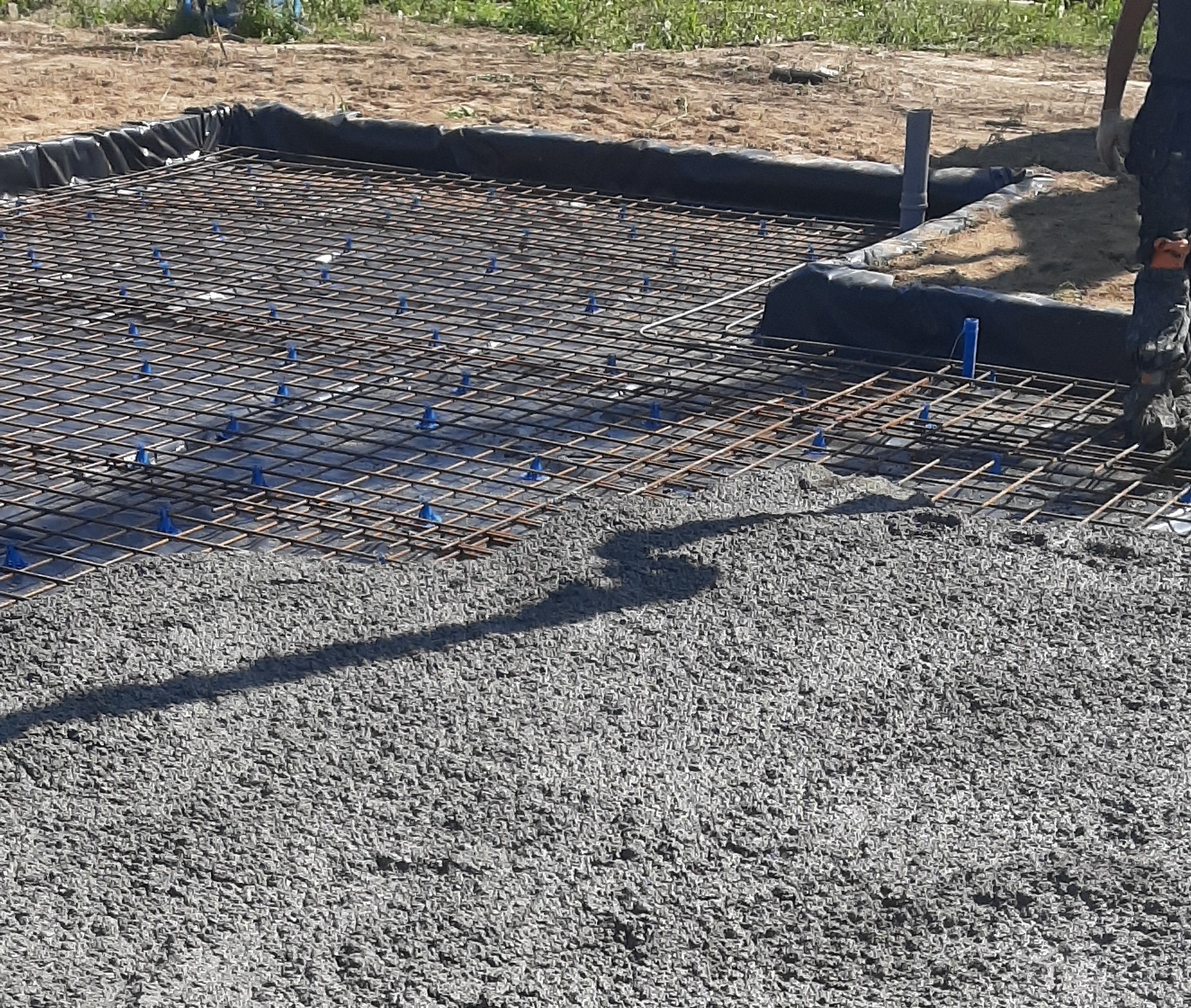 Zalewanie betonem zbrojenia na gruncie, widoczne plastikowe podkładki dystansowe pod siatką zbrojeniową i czarna folia izolacyjna na obrzeżach wykopu.