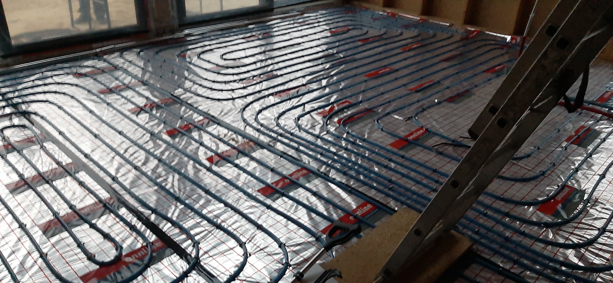 Rozłożona instalacja wodnego ogrzewania podłogowego na srebrnej folii izolacyjnej z widocznymi czerwonymi elementami mocującymi, drabina oparta o ścianę, surowy stan wykończenia.