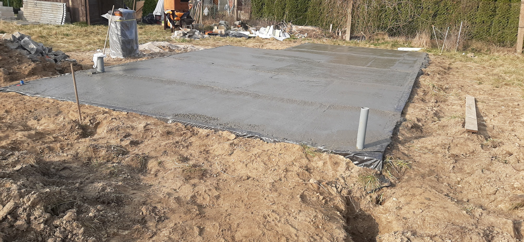 Świeżo wylana, gładka płyta fundamentowa na czarnej folii, wystające rury kanalizacyjne. W tle narzędzia budowlane, ziemia i trawa.