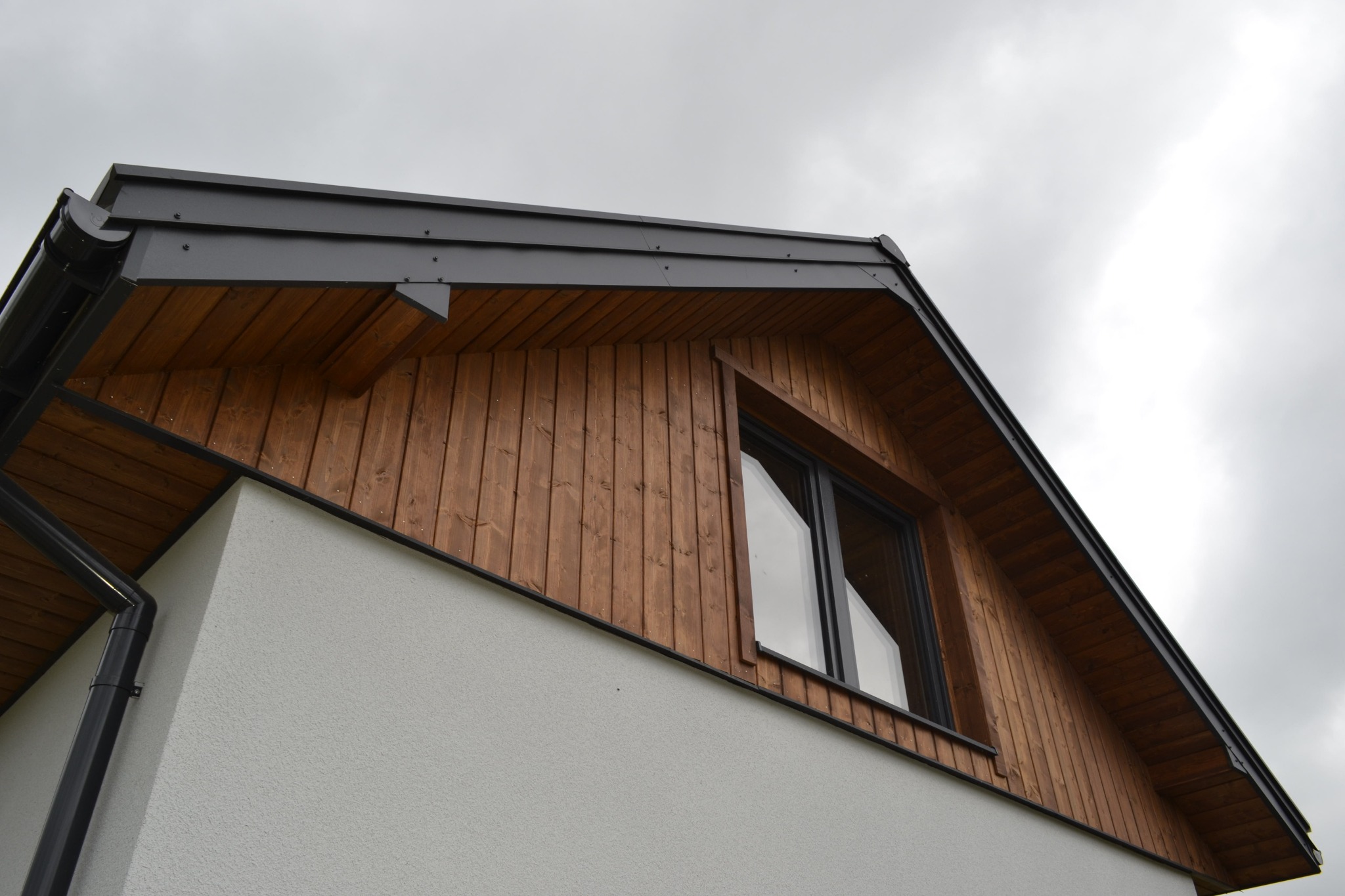 Fragment ściany zewnętrznej budynku z drewnianą elewacją, czarnym orynnowaniem i oknem w kształcie trapezu, na tle pochmurnego nieba.