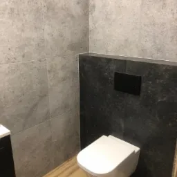 Nowoczesna łazienka po remoncie: szare płytki imitujące kamień na ścianach, podwieszana toaleta z czarnym przyciskiem spłukującym i drewnopodobna podłoga.