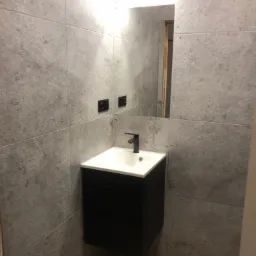 Wykończona łazienka z szarą, betonową okładziną ścienną, czarną szafką pod umywalką i drewnianą podłogą. Widoczne lustro z oświetleniem.