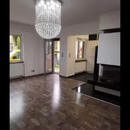 Salon po remoncie z drewnopodobną podłogą z nadrukowanymi napisami, nowoczesnym kominkiem i kryształowym żyrandolem.