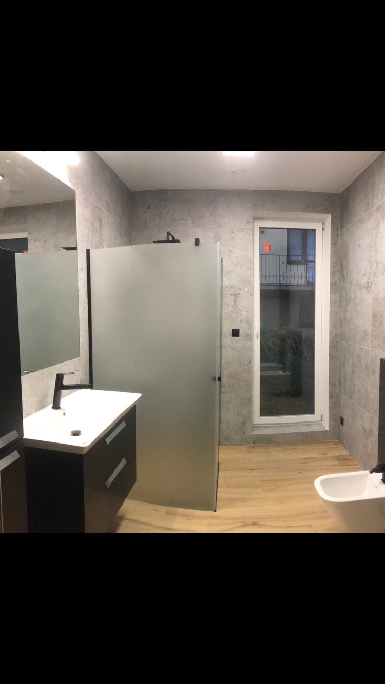 Nowoczesna łazienka z kabiną prysznicową ze szkła matowego, umywalką z szafką w kolorze ciemnego drewna, płytkami imitującymi beton i jasną drewnianą podłogą.