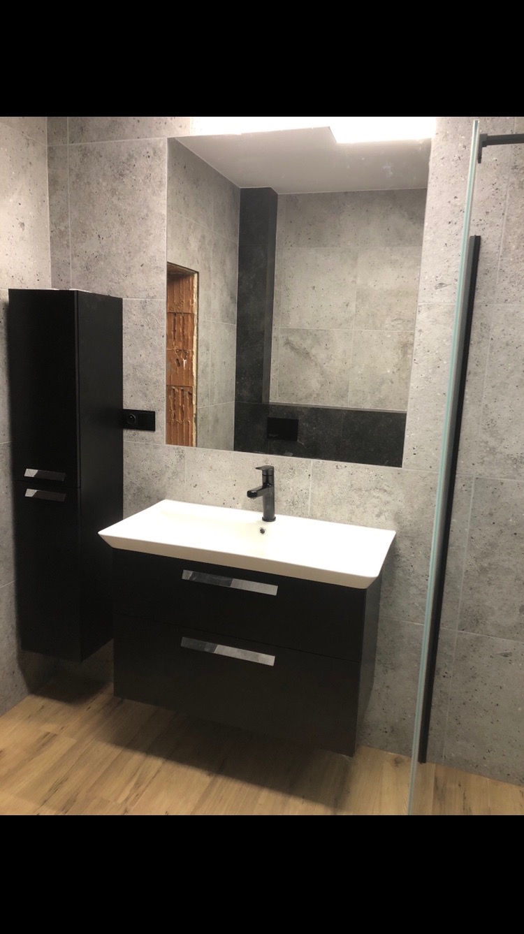 Nowoczesna łazienka z czarną szafką pod umywalką i lustrem. Szare płytki na ścianach i drewnopodobne panele na podłodze. Widoczny fragment kabiny prysznicowej.