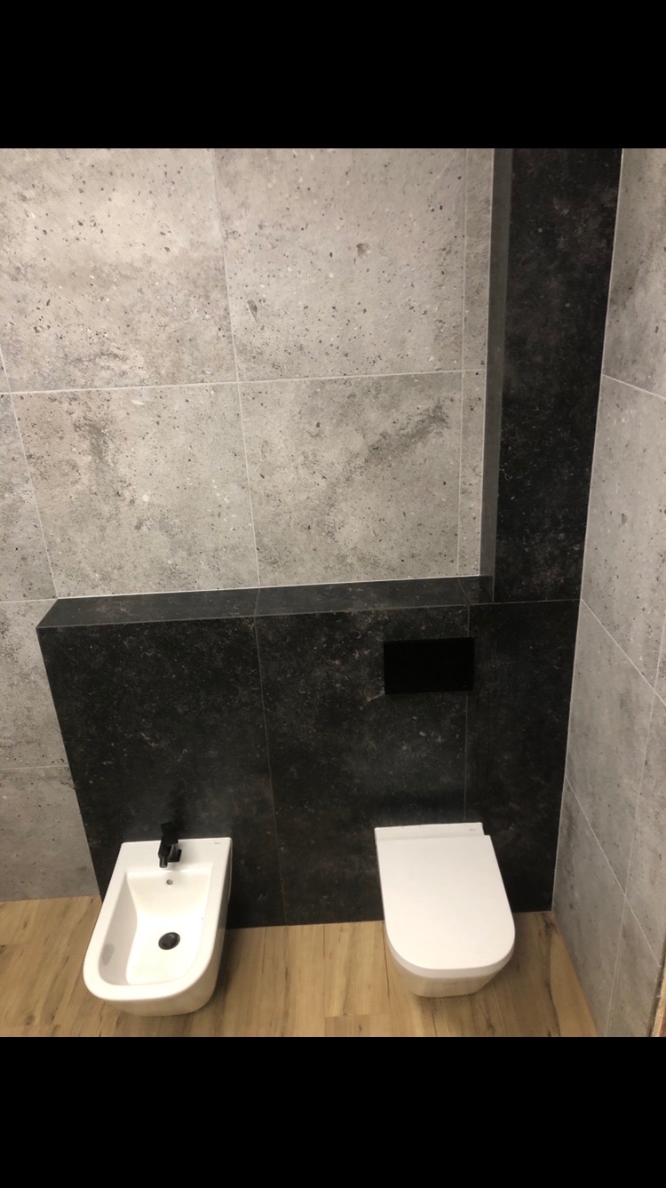 Wykończona łazienka z szarymi i czarnymi płytkami, drewnopodobną podłogą, białym bidetem z czarną armaturą i podwieszaną toaletą. Nowoczesny design.