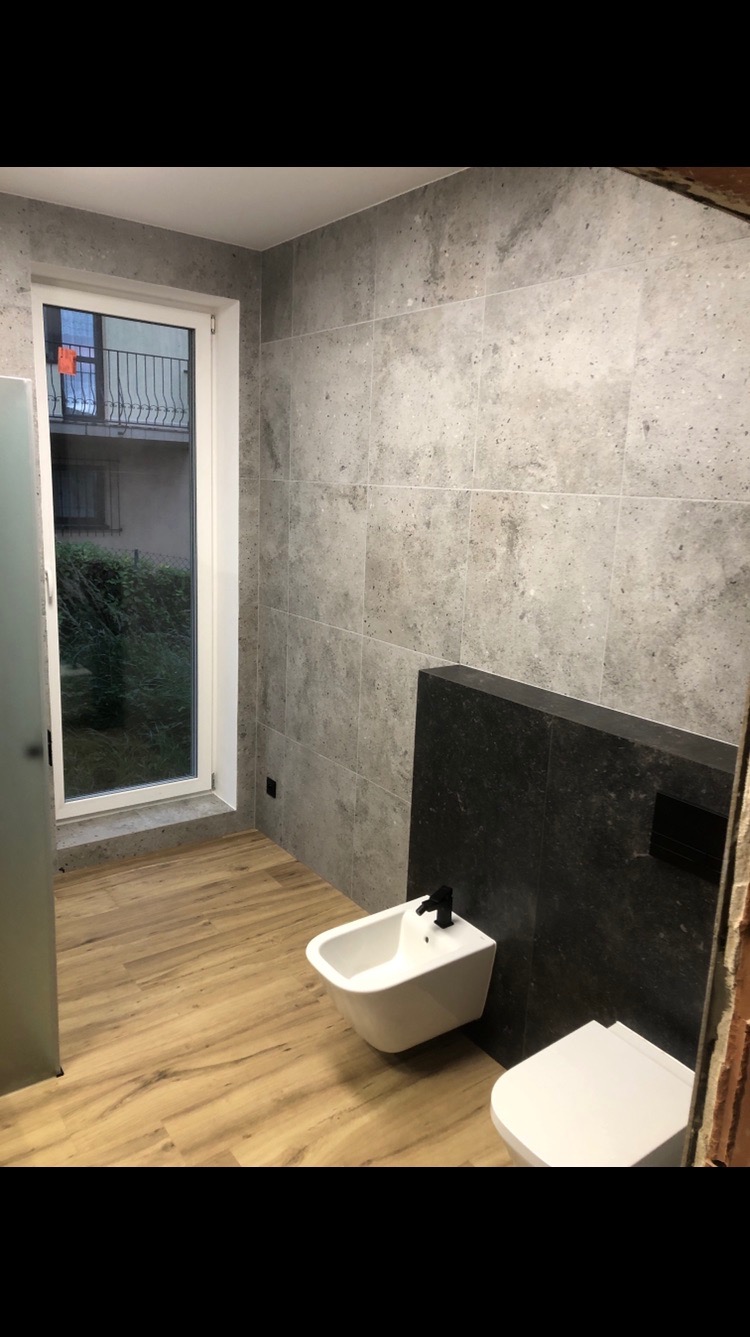 Jasna łazienka z szarymi płytkami, drewnopodobną podłogą i białymi sanitariatami. Widoczne okno z widokiem na zieleń i balkon. Minimalistyczny design.