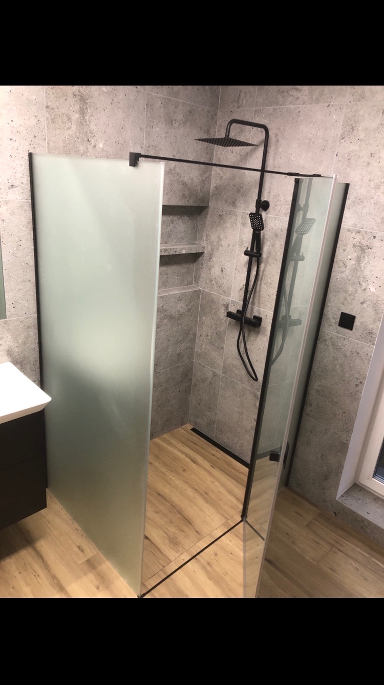 Nowoczesna kabina prysznicowa z grafitową armaturą i szklaną ścianką, wykończona płytkami imitującymi beton i drewnopodobną podłogą. W tle widoczne półki.