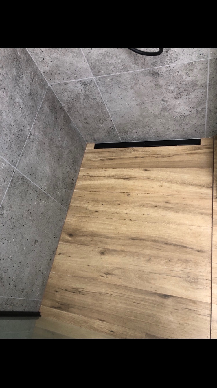 Nowoczesna łazienka z drewnopodobną podłogą i szarą, kwadratową glazurą na ścianach. Widoczny czarny odpływ liniowy.