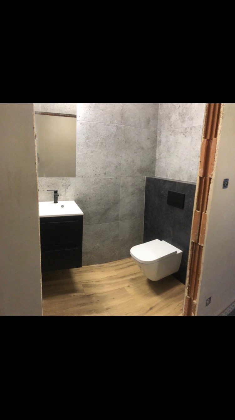 Wykończona łazienka z szarymi płytkami imitującymi beton, drewnopodobną podłogą i wiszącą toaletą. Widok zza framugi drzwi w stanie surowym.