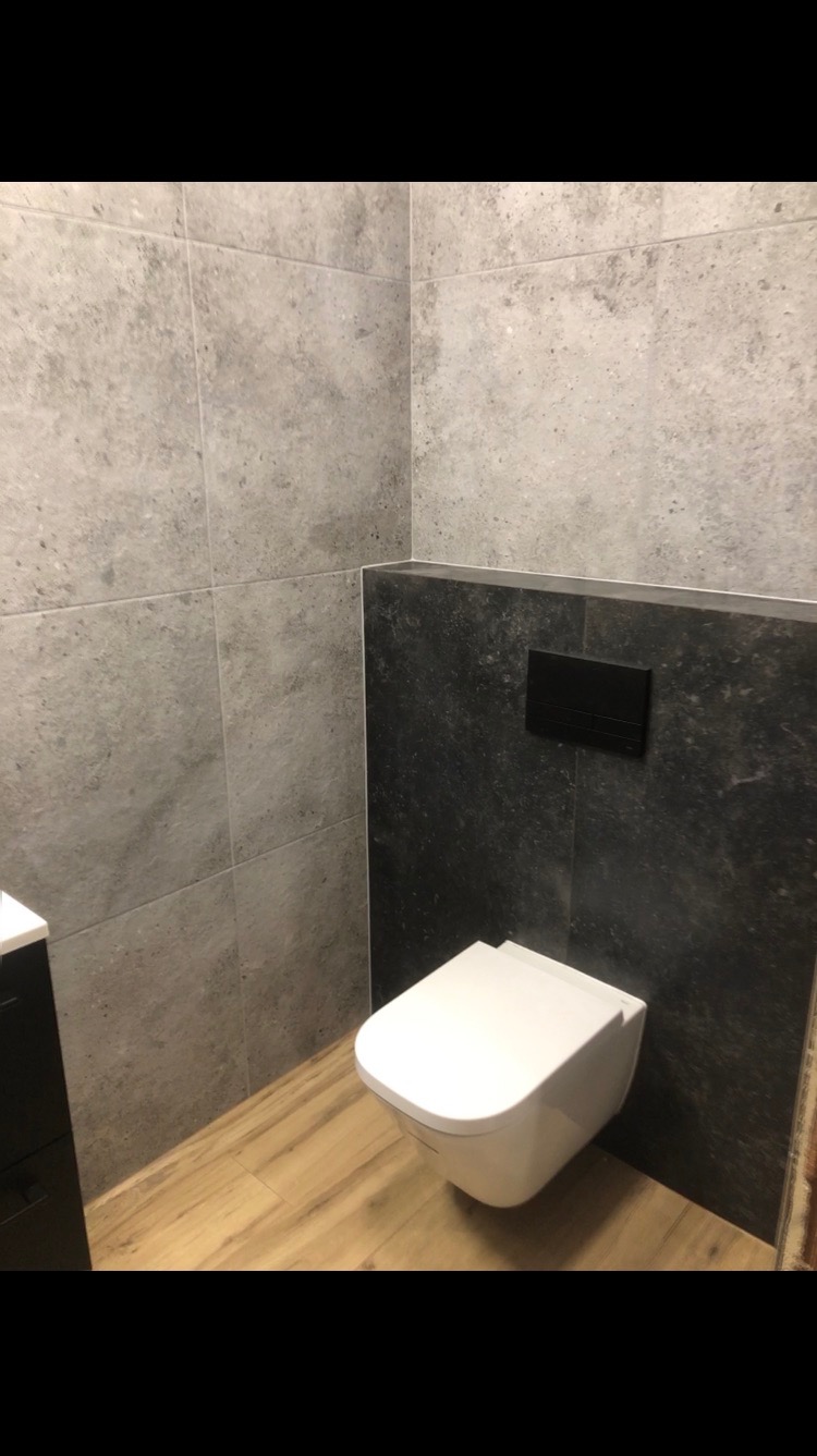Nowoczesna łazienka po remoncie: szare płytki imitujące kamień na ścianach, podwieszana toaleta z czarnym przyciskiem spłukującym i drewnopodobna podłoga.