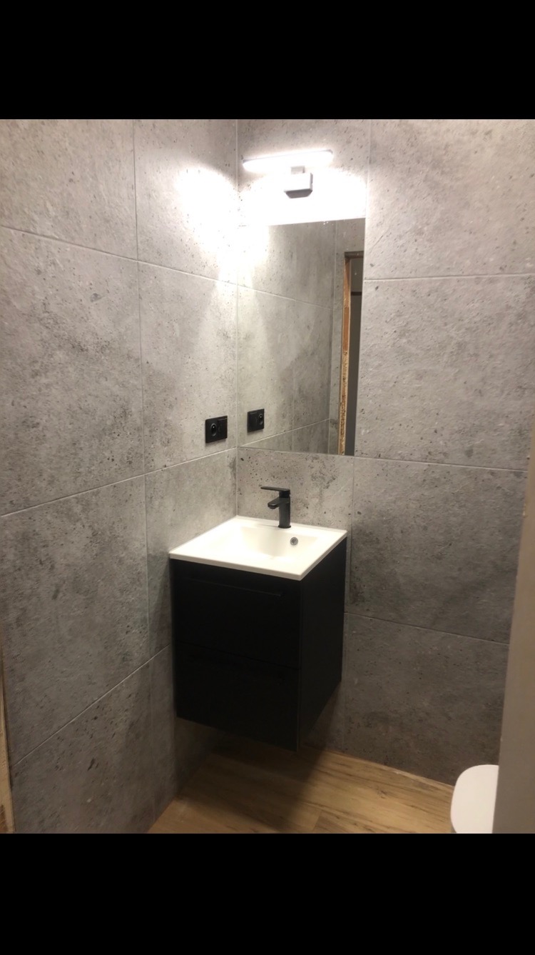 Wykończona łazienka z szarą, betonową okładziną ścienną, czarną szafką pod umywalką i drewnianą podłogą. Widoczne lustro z oświetleniem.