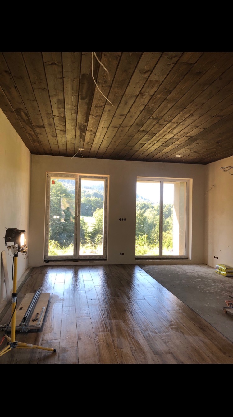 Jasne panele podłogowe imitujące drewno w nowym wnętrzu z drewnianym sufitem. Widoczny reflektor budowlany i narzędzia glazurnicze.