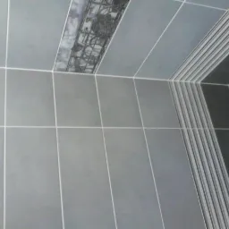 Szare płytki ceramiczne z białymi fugami na ścianie, z dekoracyjnym pasem z wzorem i pasem z pionowymi paskami.