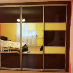 Szafa przesuwna z lustrzanymi drzwiami i panelami w kolorze ciemnego brązu oraz żółtego w sypialni, odbicie wnętrza pokoju w lustrach.