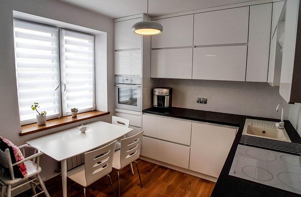 Jasna kuchnia z białymi, błyszczącymi frontami szafek, czarnym blatem i drewnianą podłogą. Nad stołem z krzesłami wisi okrągła lampa. Na parapecie okna rosną małe rośliny.