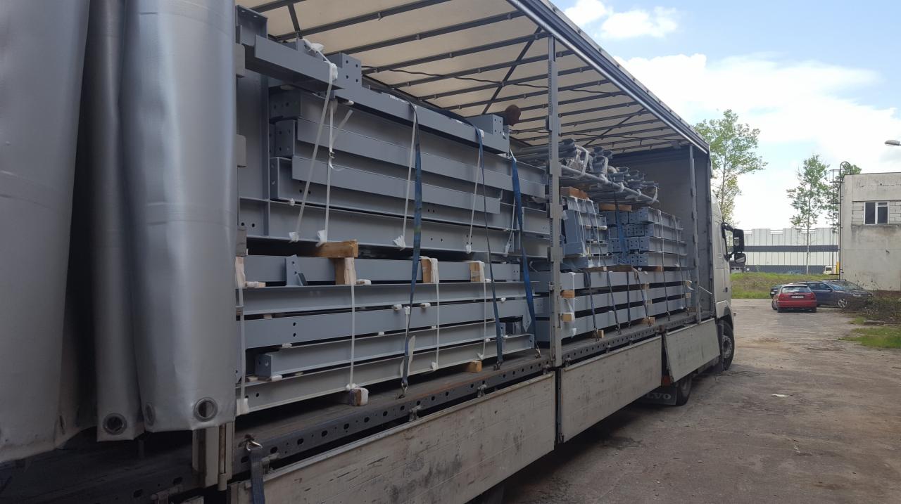 Ciężarówka z otwartą plandeką, wypełniona szarymi elementami stalowymi konstrukcji, zabezpieczonymi pasami transportowymi, widok z boku na placu budowy.