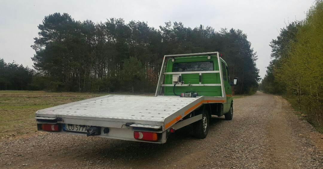 Zielona laweta z aluminiową platformą, zaparkowana na żwirowej drodze w pobliżu lasu, widok z tyłu, widoczna tablica rejestracyjna.