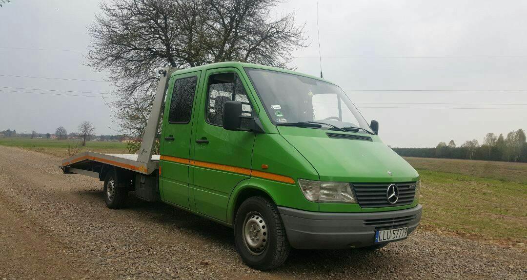 Zielona laweta Mercedes Sprinter z pomarańczowym pasem na boku, zaparkowana na żwirowej drodze polnej z widoczną tablicą rejestracyjną i drzewami w tle.