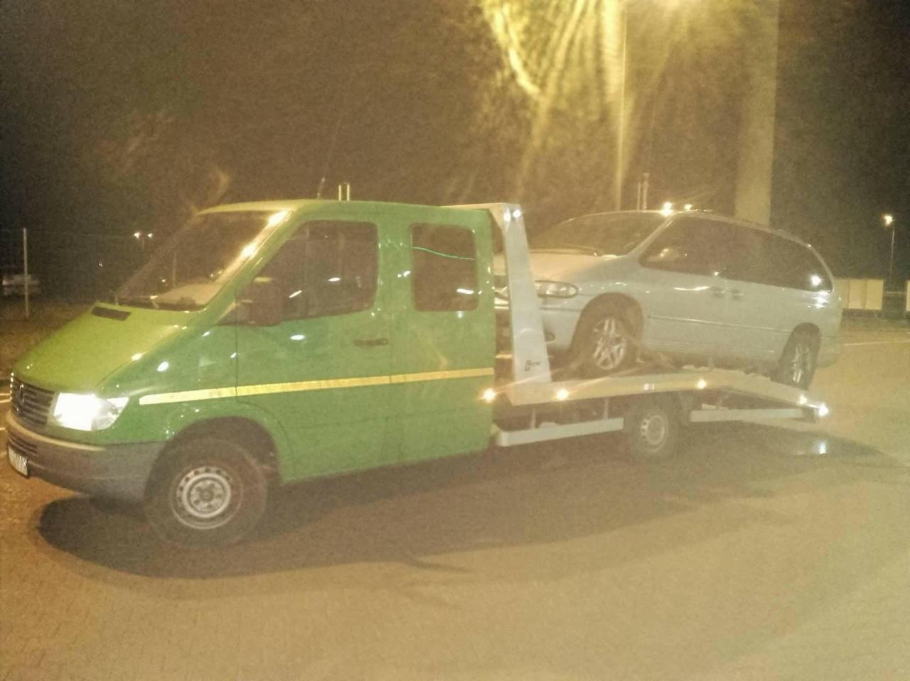 Zielona laweta Mercedes z załadowanym szarym minivanem Chrysler na parkingu oświetlonym lampami ulicznymi, widoczne mokre podłoże i słabe oświetlenie.